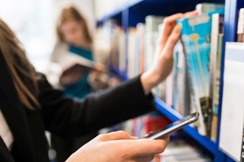 Hand of teenage girl holding cell phone while taking book from shelf in a public library
